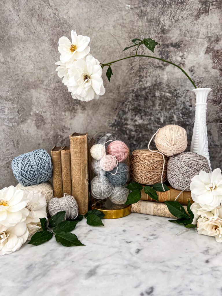 A collection of yarn, antique books, and white roses arranged in a gentle curve against a gray background. In the center is a glass cloche full of small yarn balls.