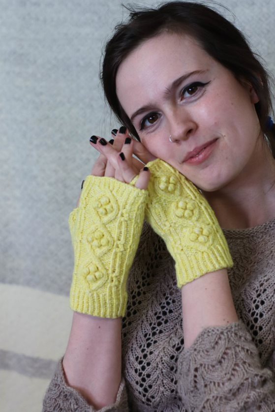 A young, white woman with dark brown hair rests her cheek against her hands, which are wearing a pair of yellow fingerless mitts with cables and bobbles running up the back.