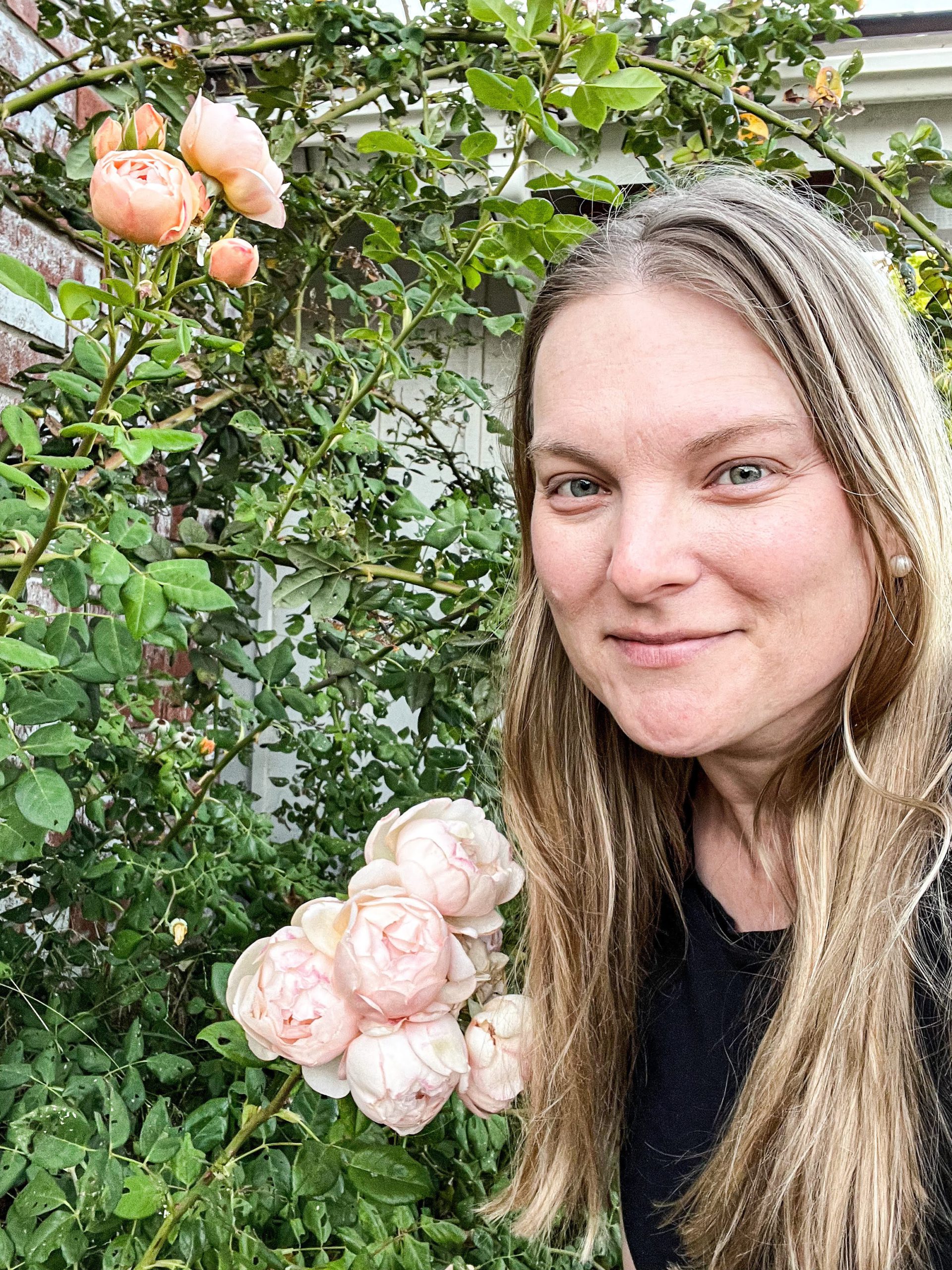 A blonde, white woman with long hair smiles with her mouth closed next to a few clusters of peach roses.