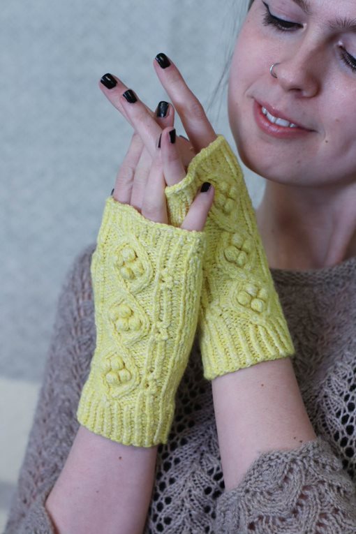 A young, white woman smiles softly at her hands, which are wearing a pair of yellow, fingerless mitts with cables and bobbles running up the back.