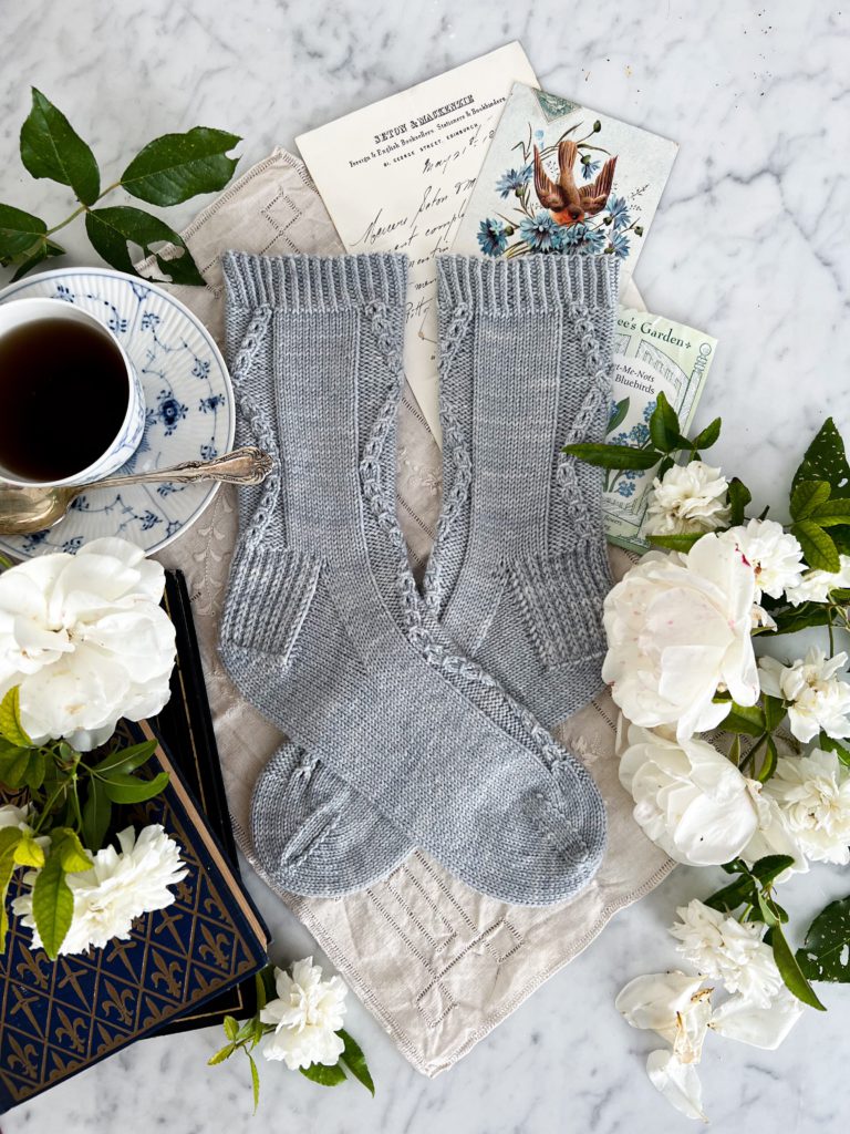 The Deimatic Socks, a pair of light blue, cabled socks are laid flat on a white marble countertop. Surrounding them are white roses, antique paper ephemera, old books, and a blue and white coffee cup.