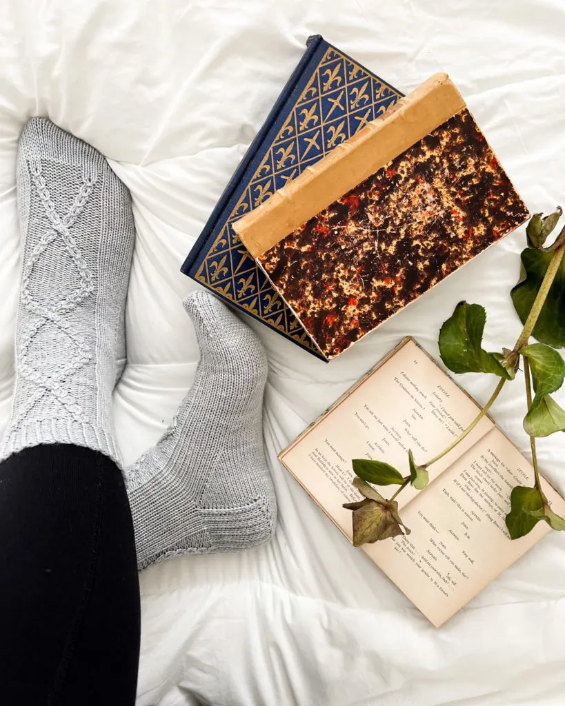 The Deimatic Socks, a pair of light blue, cabled socks on feet that are crossed at the ankle. Next to them are some antique books.