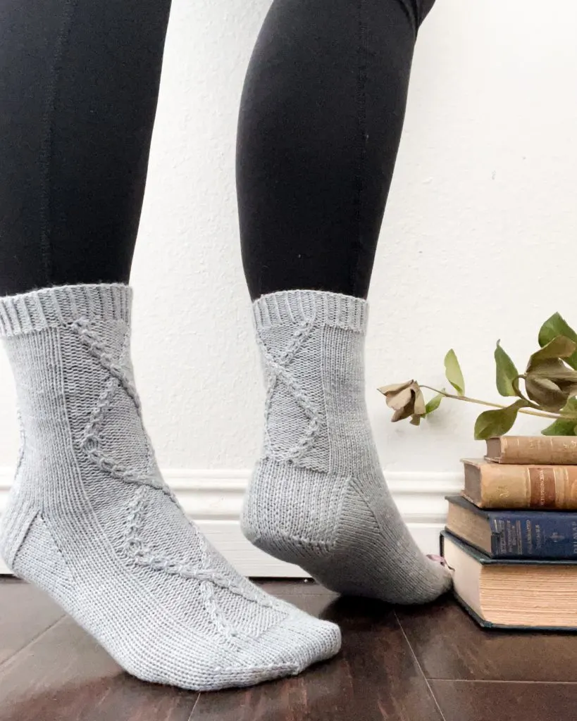 The Deimatic Socks, a pair of light blue, cabled socks. They are worn on two feet standing slightly on tiptoe. Next to them are some antique books.