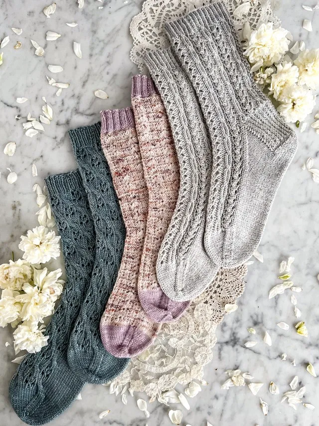 Three pairs of hand knit socks are laid out flat on a countertop. They ascend to the right in a stairstep fashion. Each pair has an all-over lacy texture.