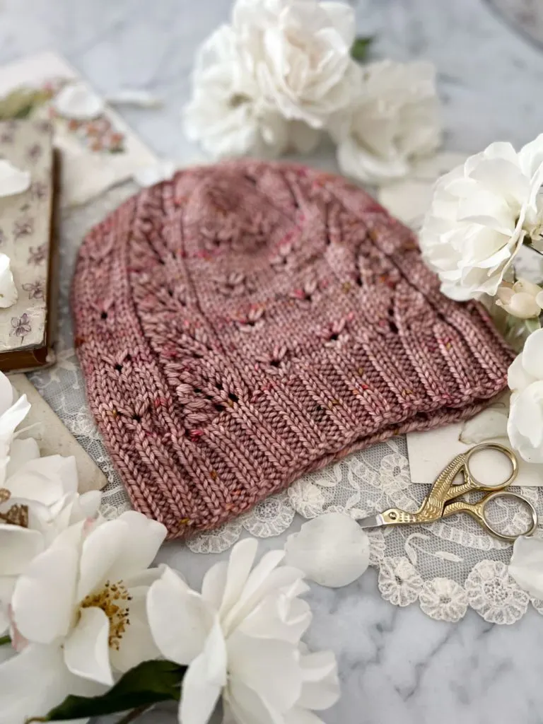 A zoomed-in image of a pink hat with lace panels and eyelets that is laid flat on a white marble countertop. The image focuses on the brim and the beginning of a lace panel. The hat is surrounded by white roses, antique paper ephemera, and an old book with a gilded cover.