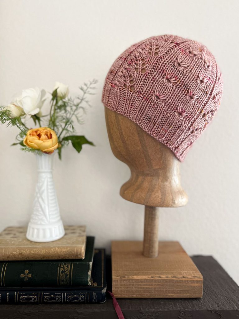 The Profiterole Hat, a pink hat with lace panels and eyelets, is shown displayed on a brown mannequin head form.