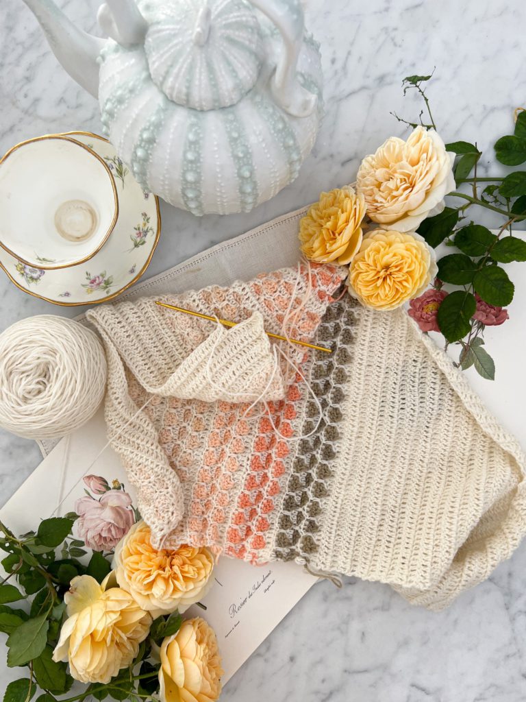 A crocheted shawl in shades of cream, green, and several pink varieties is laid out, slightly rumpled, on a white marble surface. It's surrounded by a teapot with a texture that looks like a sea urchin, an empty teacup, several yellow roses, and a couple antique rose botanical prints.