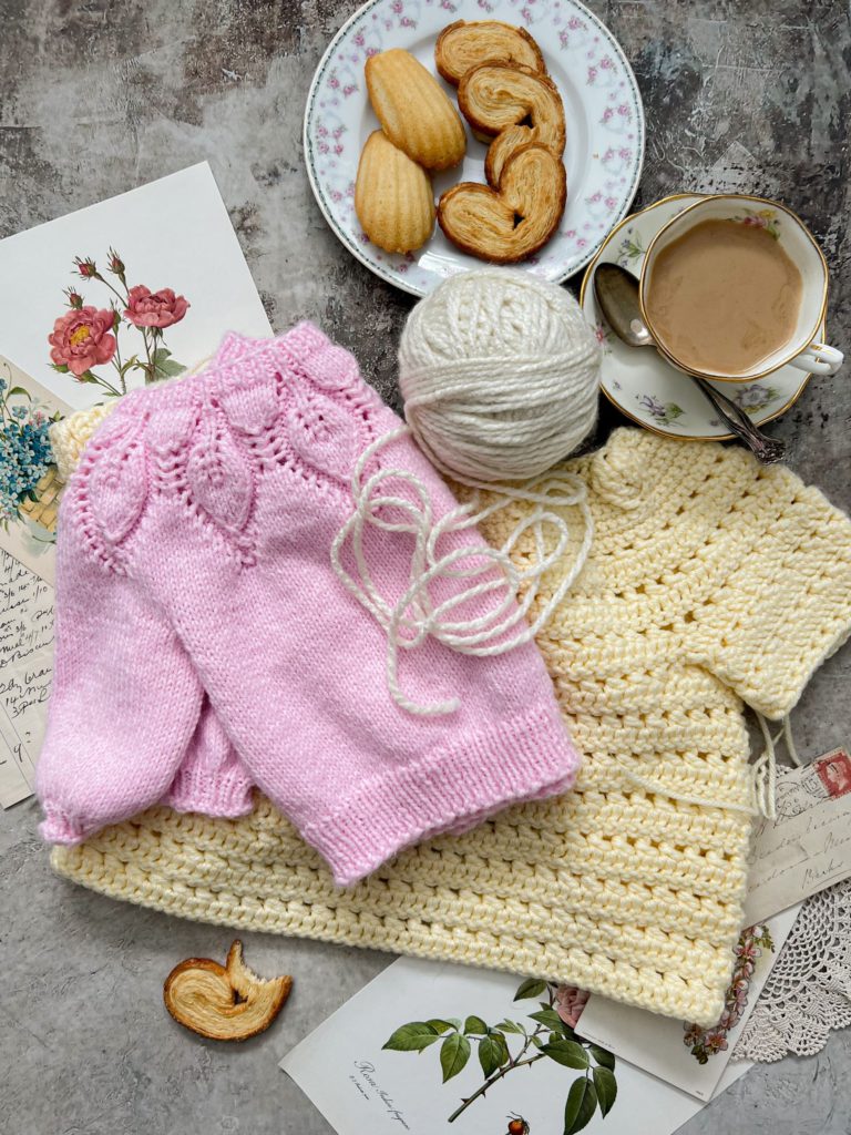 A top-down image of a pale yellow crocheted child's sweater with a pink knit child's sweater folded on top and a cream-colored ball of yarn. They're surrounded by a teacup full of coffee, a plate with tiny baked treats, and antique paper ephemera.