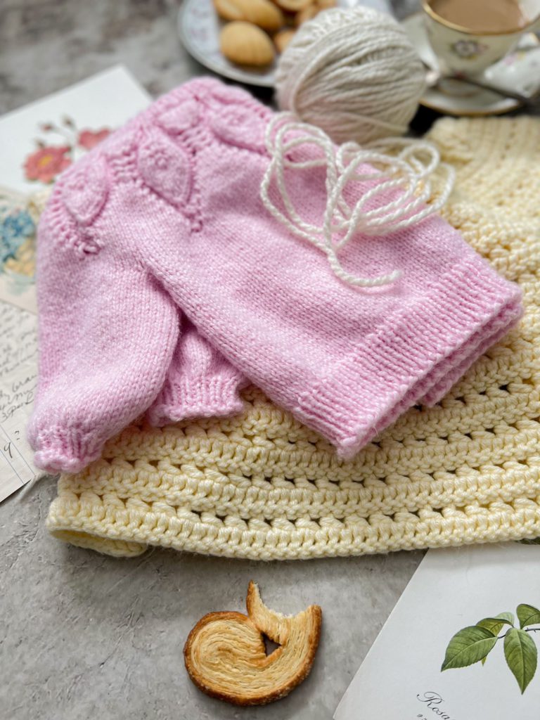 A zoomed-in image of a pale yellow crocheted child's sweater with a pink knit child's sweater folded on top. In the background is a cream-colored ball of yarn. They're surrounded by a teacup full of coffee, a plate with tiny baked treats, and antique paper ephemera.