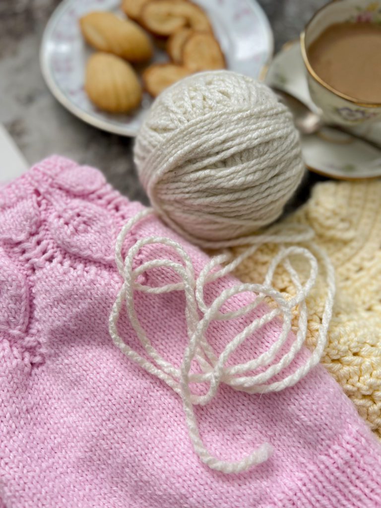 A zoomed-in image of a cream-colored ball of yarn in the center of the photo. It is surrounded by a pink knit sweater, a pale yellow crocheted sweater, and, blurred in the background, a teacup of coffee and a plate of tiny baked treats.