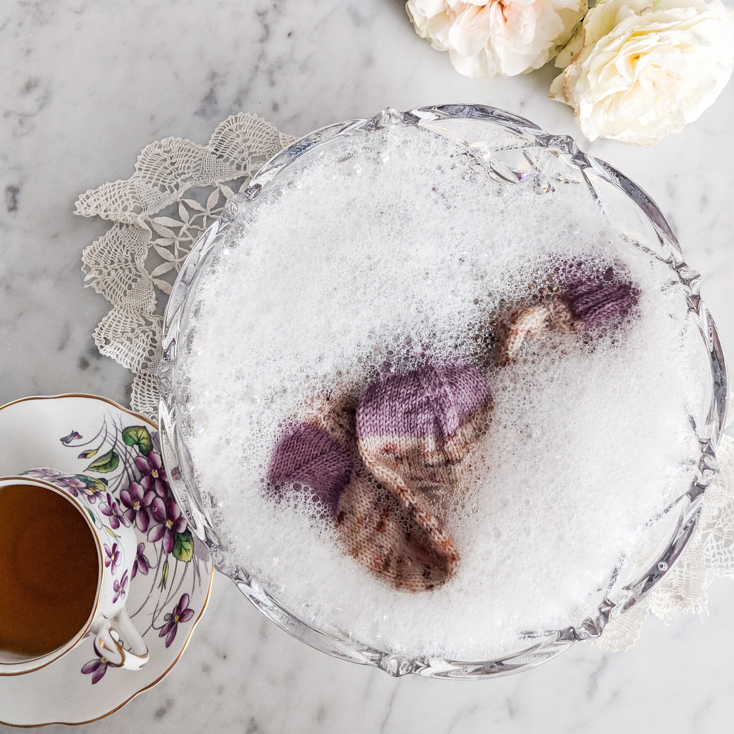 A speckled sock with purple cuffs and heels peeks through the white foam at the top of a crystal bowl. The socks are soaking as part of the blocking process.