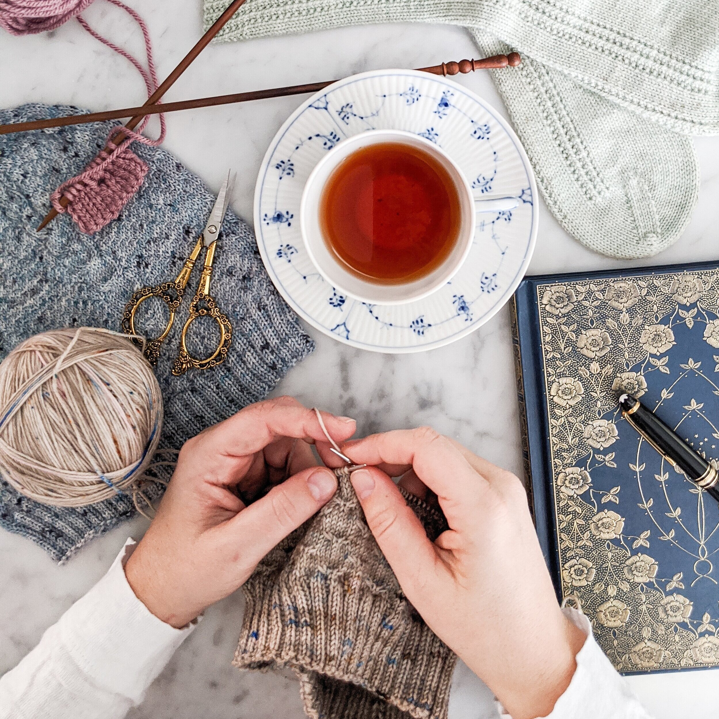 A pair of white hands works on a hat knit with light brown yarn speckled with pink and blue. They are surrounded by other knit items, a teacup filled with tea, a journal with a gilded cover, and ornate scissors. This beginner's guide to knitting will help you get started on your knitting journey.