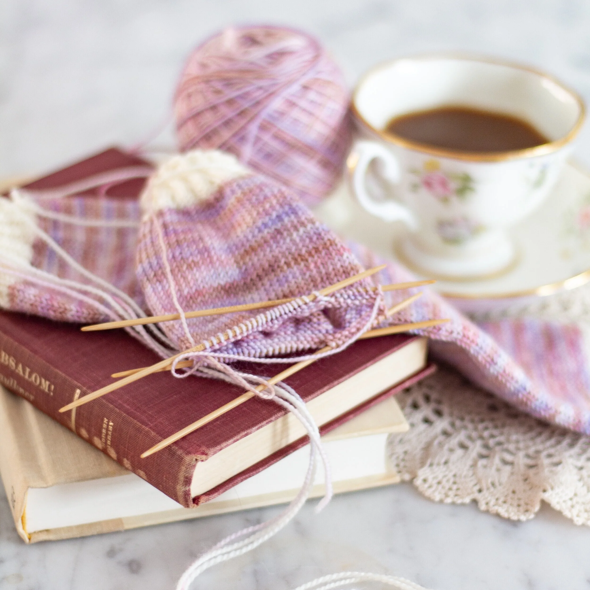 A partially-finished pair of variegated pink and white socks sit on top of a small stack of books. A teacup is slightly blurred in the background. Yarn substitution helped me learn how to use different yarn for socks, sweaters, hats, and more.