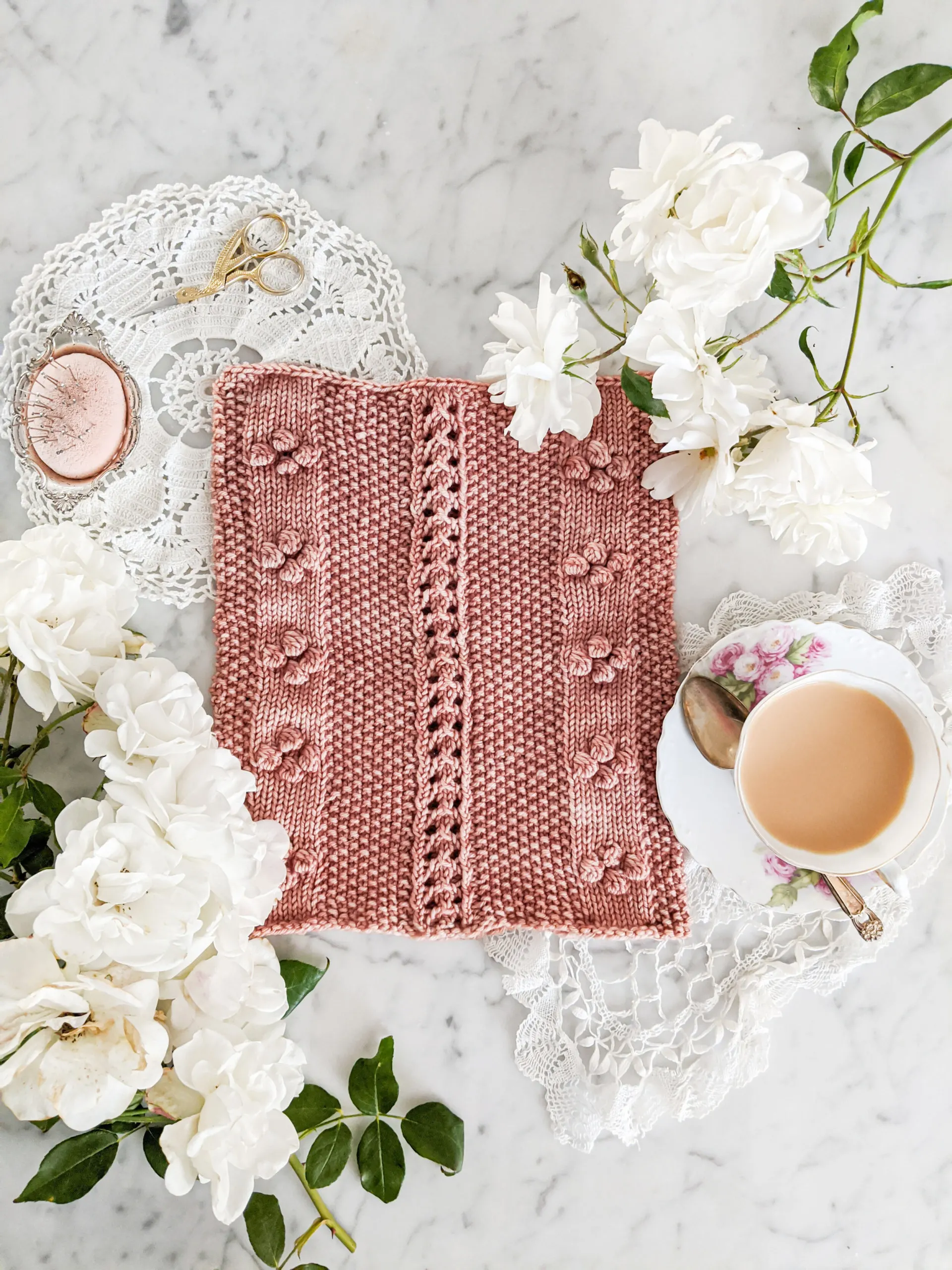 A pink cowl with columns of eyelets and bobbles is laid flat on a white marble countertop and surrounded by white roses, a teacup full of milky tea, and antique doilies.