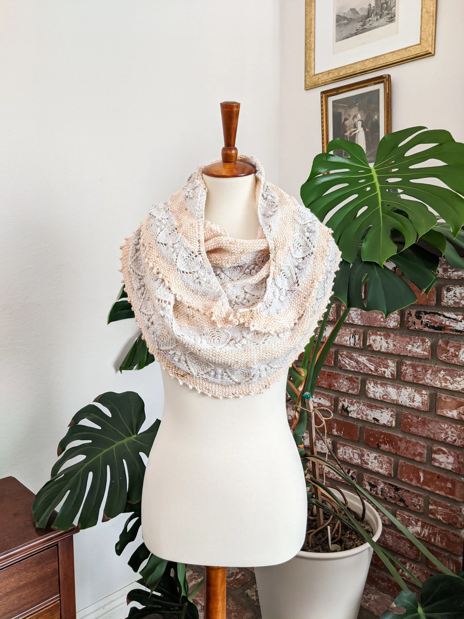 A crescent-shaped shawl in pale blue and yellow stripes is draped around the torso of a white dressmaker's form. In the background is a large monstera plant, a brick fireplace, and some framed antique prints on the walls.