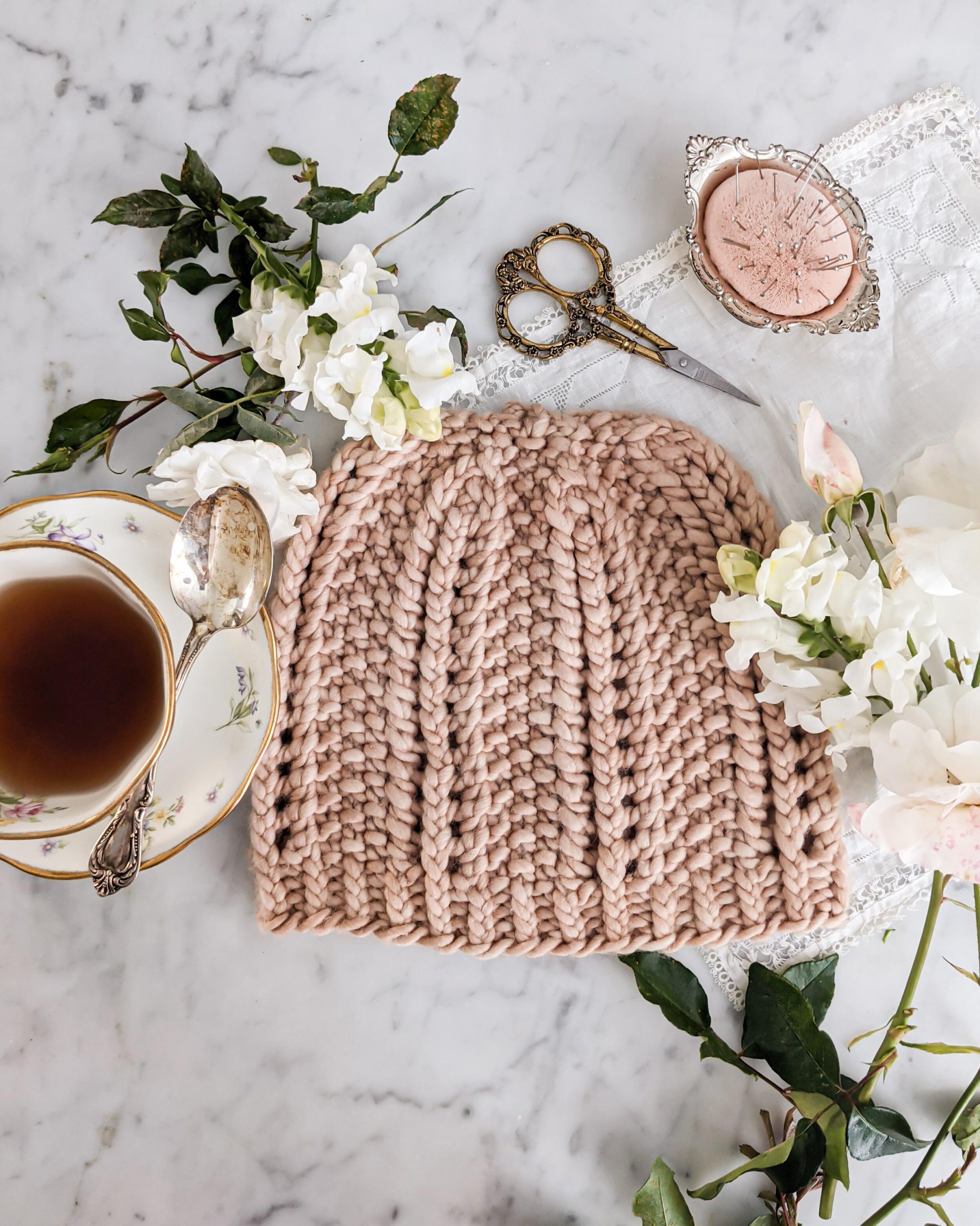 A pink hat with columns of eyelets and slipped stitches is laid flat on a countertop surrounded by a teacup, white flowers, and ornate brass scissors.