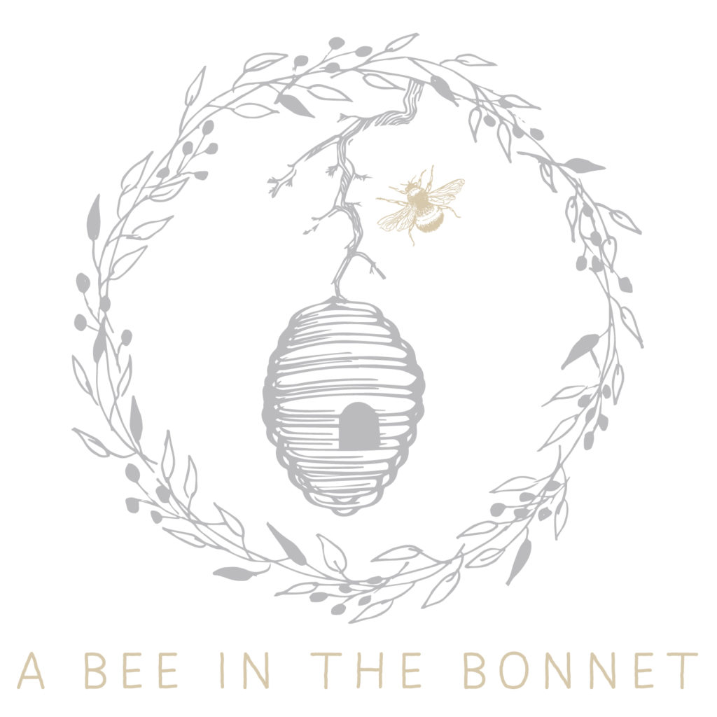 A Bee In The Bonnet Logo - A gray beehive hanging from a branch surrounded by a gray wreath of leaves and berries. A little golden bee flies toward the hive. At the bottom are the words "A Bee In The Bonnet" in gold letters.