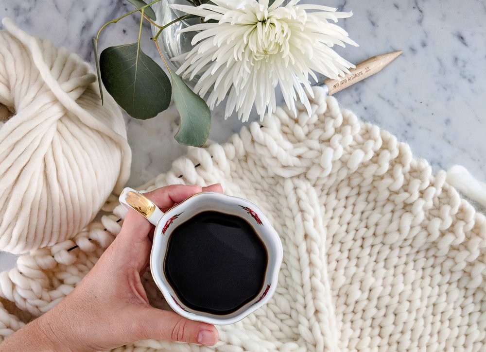 A white woman's left hand holds a mug of black coffee on top of a cream-colored knit blanket in progress. To the top left is more yarn and a large white dahlia blossom.