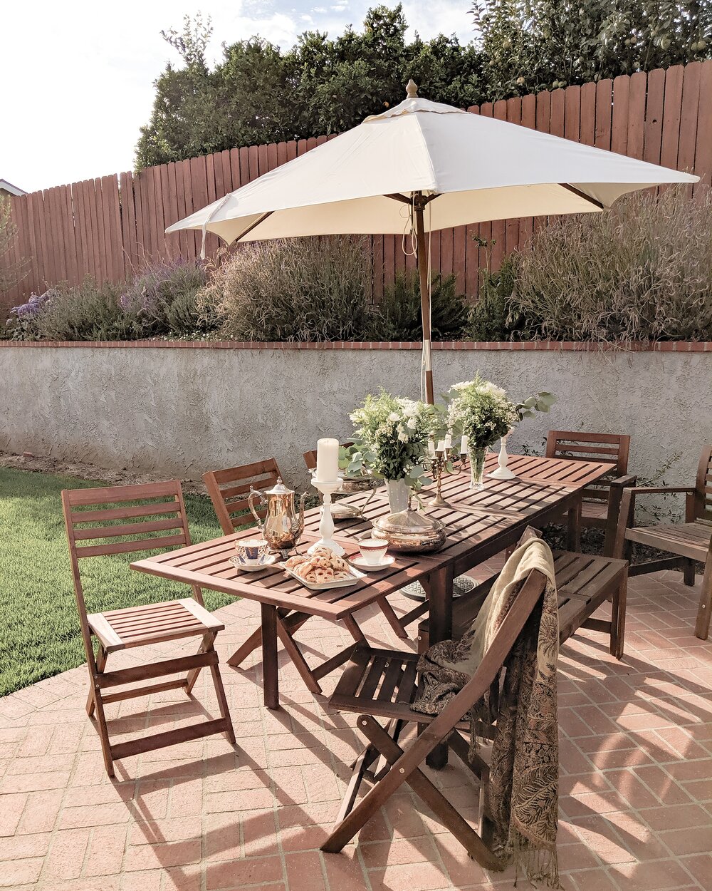 A brown wood patio set with a white umbrella on a brick patio. The table is filled with tea party supplies and vases of white flowers.