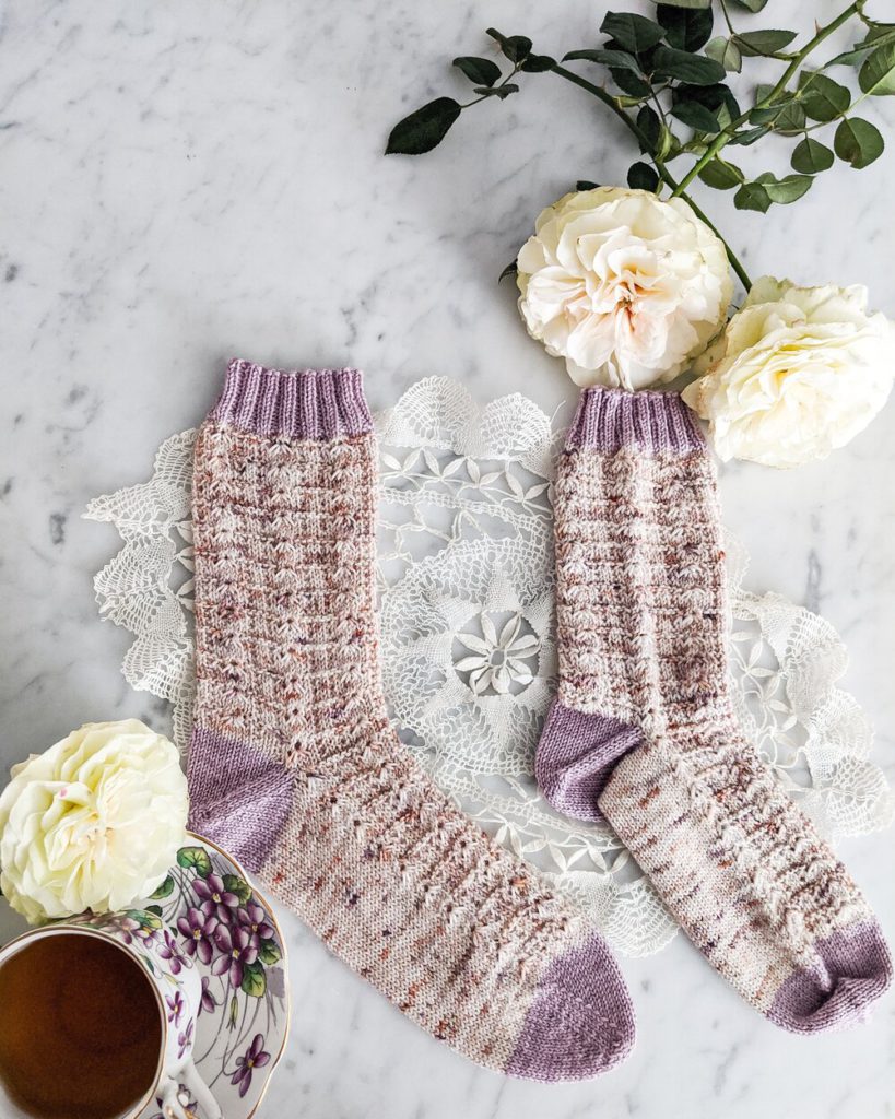 Two speckled pink and cream socks with contrasting purple cuffs, heels, and toes are laid out flat on a white marble countertop with their toes pointing to the right. The sock on the left has been blocked and is smooth. The sock on the right has not been blocked and is rumpled and scrunched.