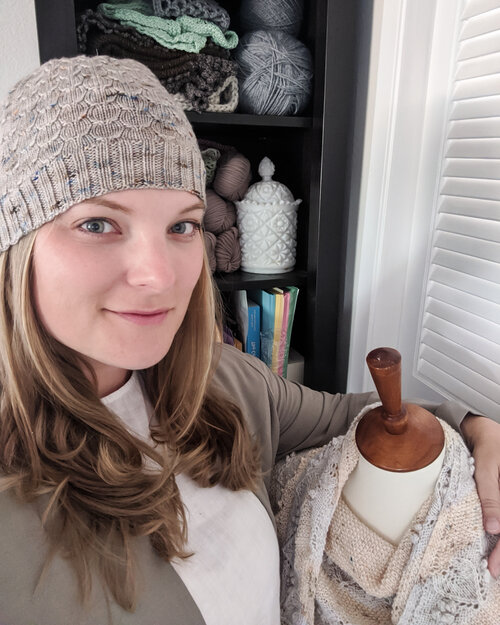 A blonde woman in her 30s wearing a knit hat, a white shirt, and a tan jacket smiles softly with her arm draped around her dressmaker's form.