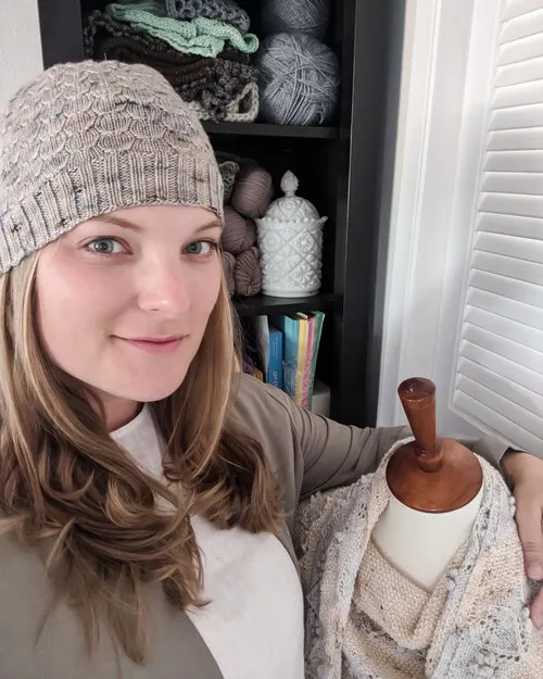 A blonde woman in her 30s wearing a knit hat, a white shirt, and a tan jacket smiles softly with her arm draped around her dressmaker's form.