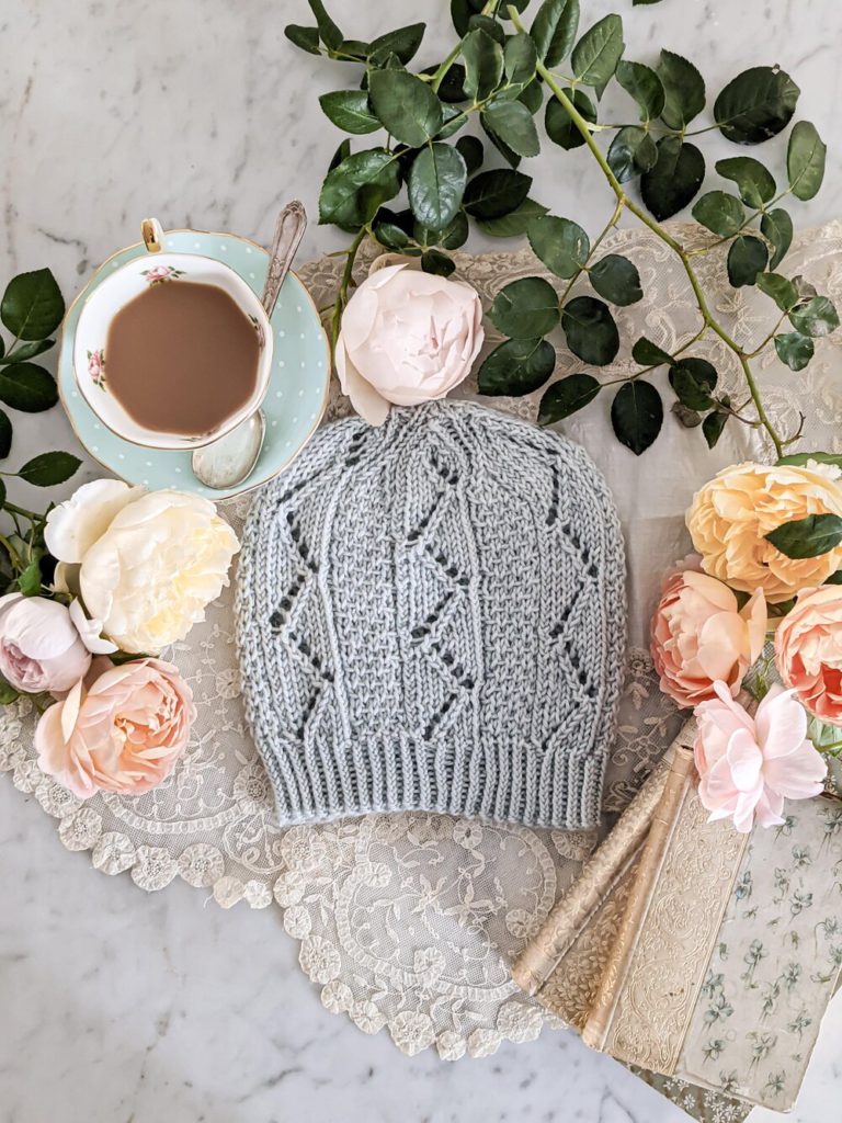 A pale blue hat with zig-zag eyelets is laid flat on a white marble countertop surrounded by pink, yellow, and orange roses, a teacup filled with coffee, and antique books.