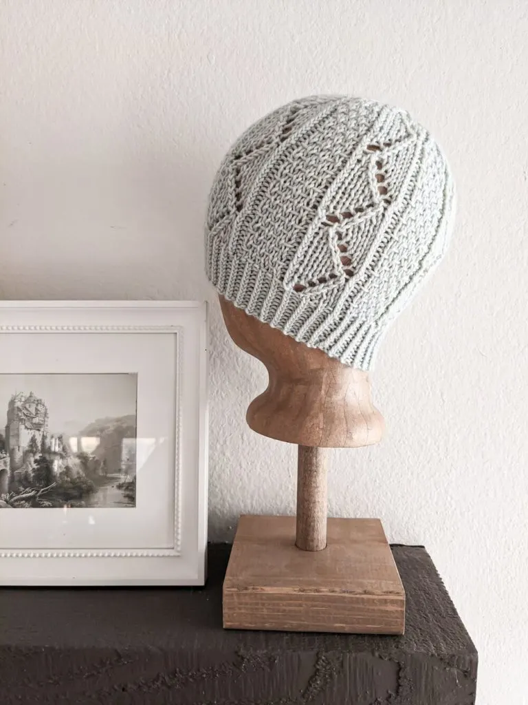 The Snuggery Hat, a light blue knit hat with zig-zag eyelets, is shown on a wooden head model. To the left is an antique print in a white frame.