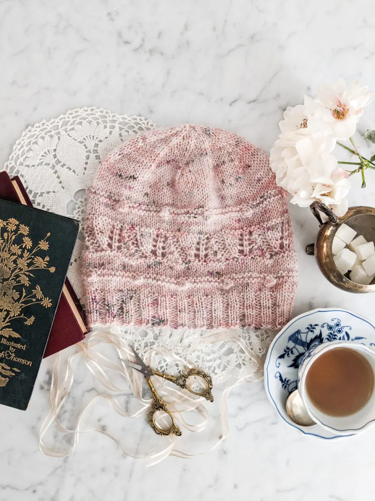 A pink hat with a horizontal band of simple lace and bobbles is laid flat on a white marble countertop. It's surrounded by antique books, white roses, and a blue and white teacup full of chamomile.
