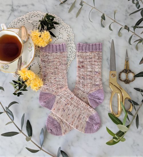 A pair of speckled pink socks with lavender cuff, heel, and toe sits on a white marble countertop with a teacup, yellow flowers, and two pairs of brass scissors. These are the Meringue Socks, the first project my tech editor Bonnie Davis and I worked on together.