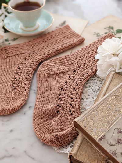 A pair of pink, lacy socks are laid out on a white surface with antique books, white roses, and a pale green teacup. My test knitters helped me get this pattern ready for publication, and I didn't have to impose any strict requirements.