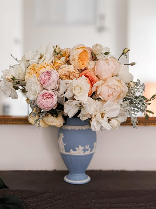 A pale blue Wedgwood Jasperware vase holds a collection of pink, peach, and yellow David Austin roses along with white iceberg roses and dusty miller.