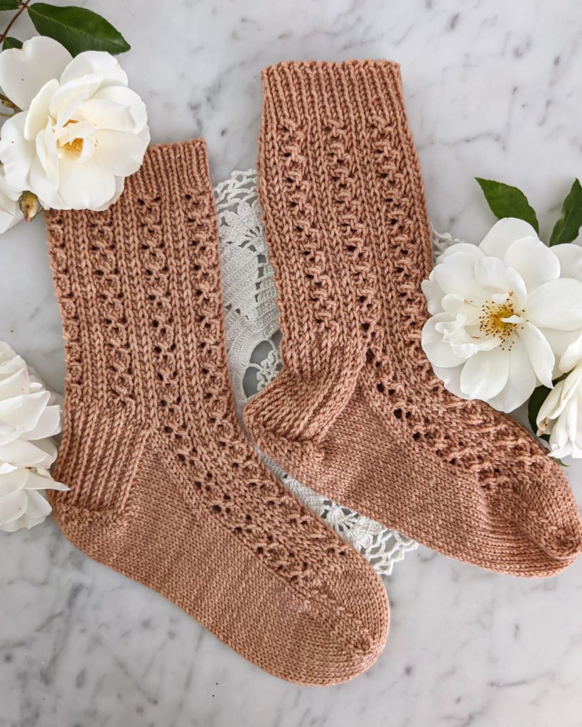 Two knit socks in a clay-colored yarn are laid flat on a white marble counter. One has been blocked and one hasn't.