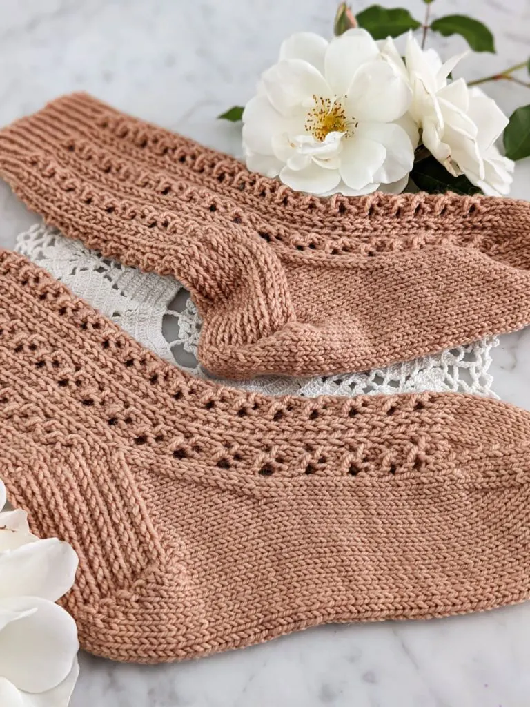 A close-up on the heels of two clay-colored, lacy knit socks. One has been blocked, and therefore sits smooth and flat. The other had not been blocked, and is therefore a bit lumpy.