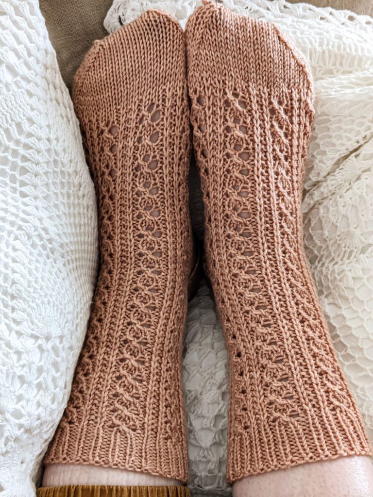 Two lacy, clay-colored knit socks on a pair of feet, resting on white crocheted pillows. The sock on the left sits smoothly on the foot, while the sock on the right is a little wrinkled and puckered.