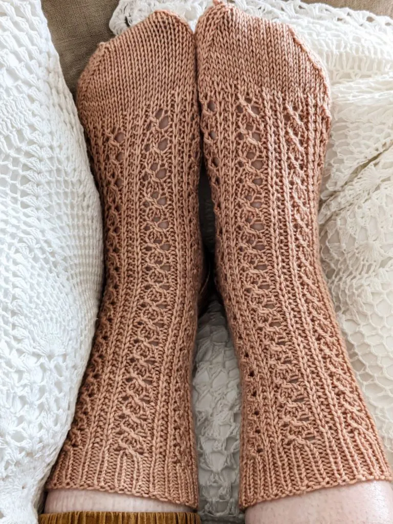 Two lacy, clay-colored knit socks on a pair of feet, resting on white crocheted pillows. The sock on the left sits smoothly on the foot, while the sock on the right is a little wrinkled and puckered.