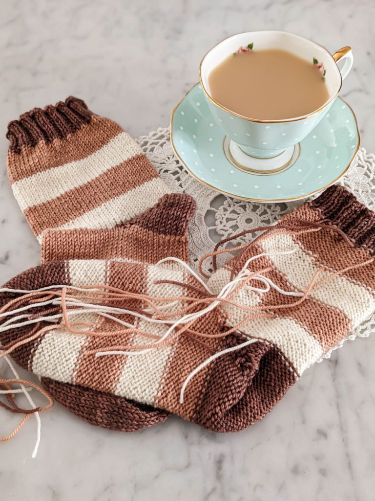 Two handknit striped socks in pink, white, and brown are laid out on a white marble countertop with a mint green teacup and saucer. One sock is turned inside out to show all the loose ends from the stripes. Weaving in ends on knitting projects like this is tedious, but the clasped weft join can help avoid that issue.