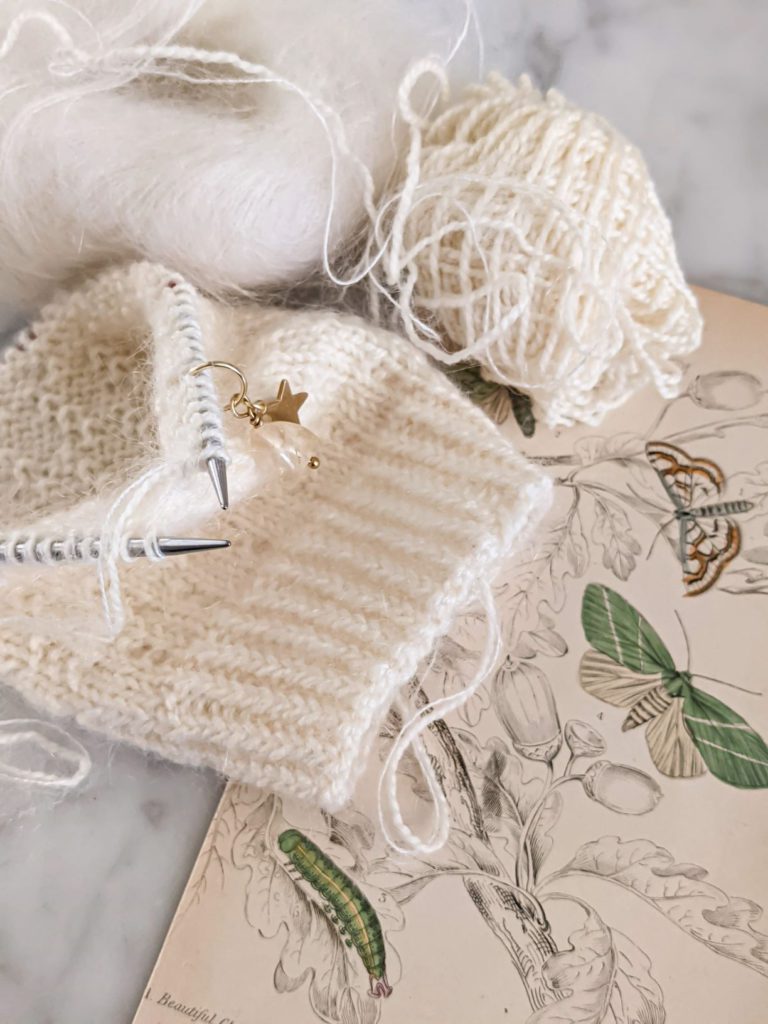 A small, white sock on tiny circular needles is sitting on a white marble countertop with an antique botanical print and two balls of white yarn. These tiny circular needles are some of my favorite needles for knitting socks.