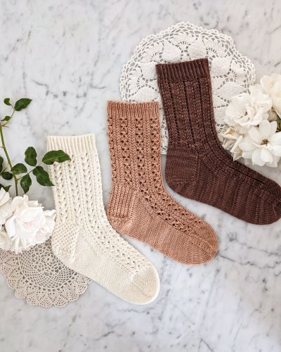 Three lacy socks - one white, one pink, and one brown - are laid flat on a white marble countertop with two doilies and some white roses. These are the Bricolage Socks, the Cromulent Socks, and the Parnassian Socks.