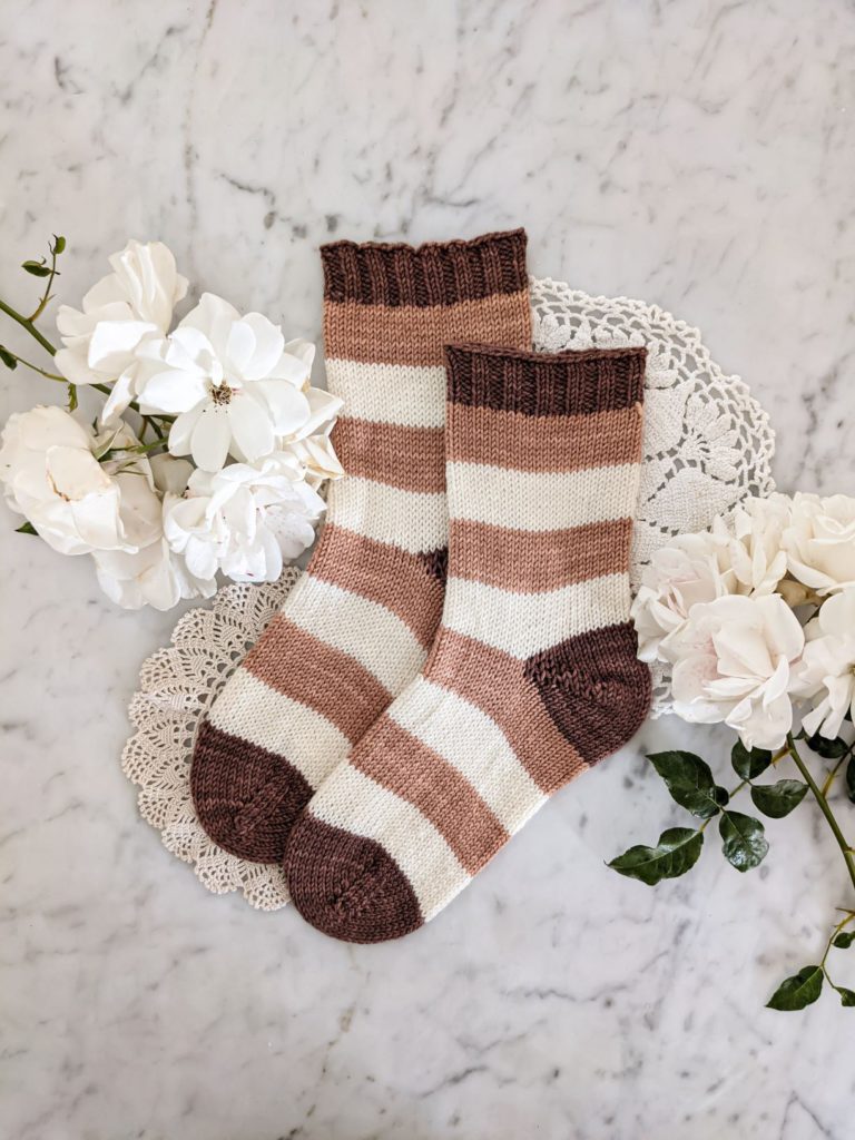 A pair of pink, white, and brown-striped socks are laid flat on a white marble surface with doilies and white roses. I had to experiment to find the right bind-off for my toe-up socks.