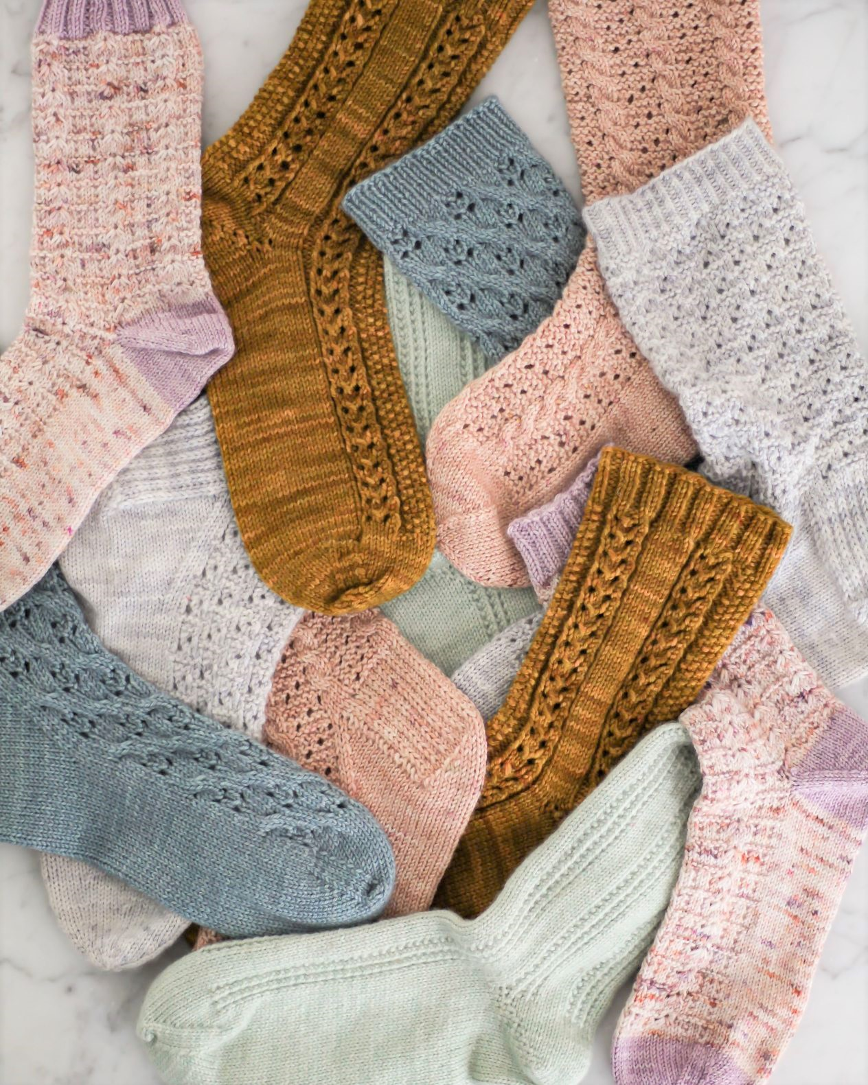 A pile of pastel handknit socks all laid out on a flat surface. There are pink, blue, light green, and caramel-colored socks jumbled up together.