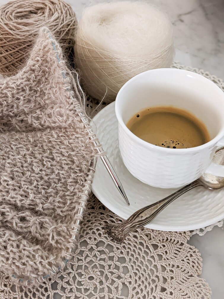 A white teacup half filled with coffee sits on a white saucer with a partially knit hat next to it. The hat is made with one strand of tan wool and one strand of white silk-mohair. I did a lot of knitflixing while working on this project.