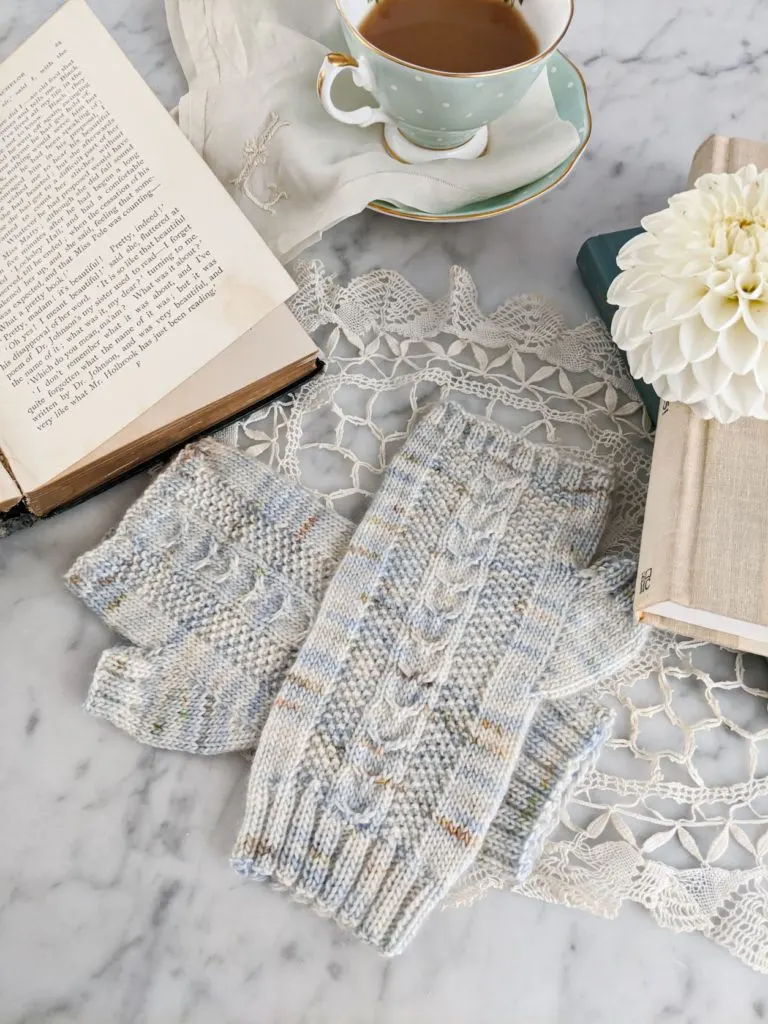 A pair of pastel fingerless mitts are laid flat on a white marble countertop, surrounded by books, a teacup, and a dahlia. The small mitts aren't overwhelmed because the larger props have been cropped. This is part of why framing your photograph is so important.
