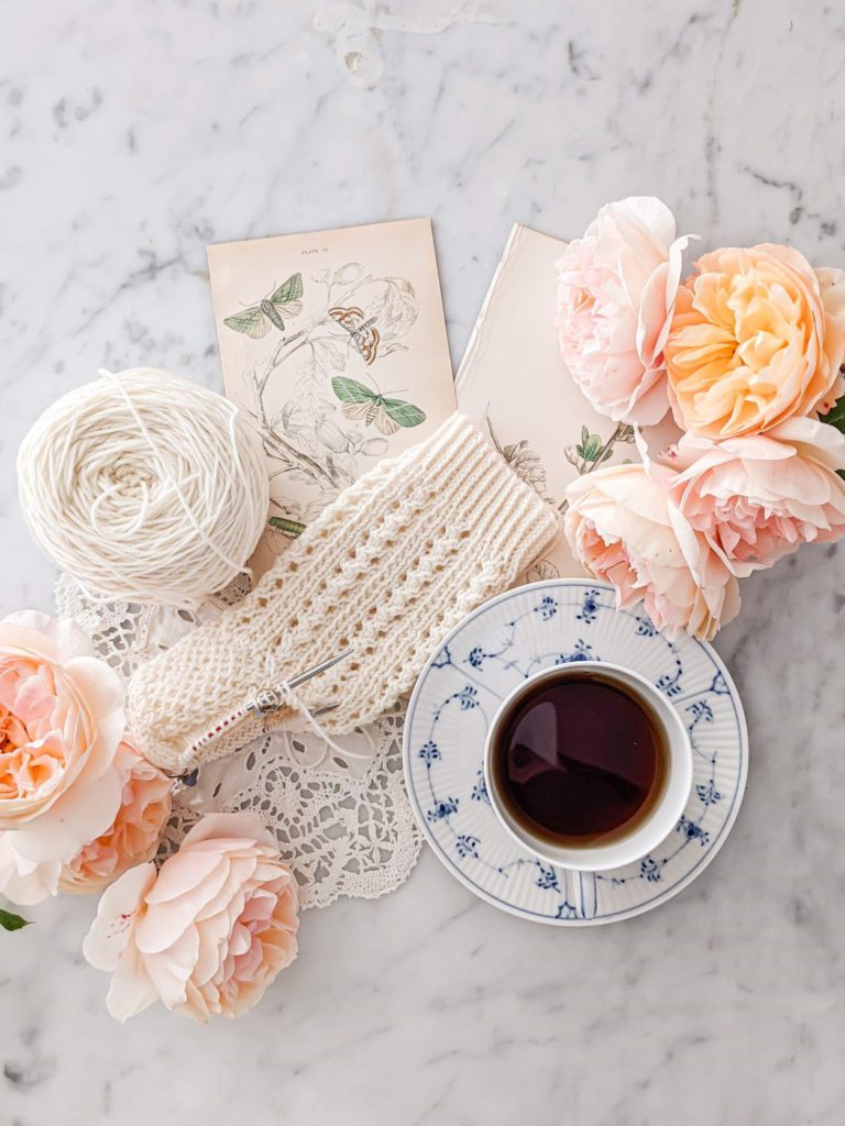 A partially-knit white sock sits on a white marble countertop with antique botanical prints, a doily, peachy-pink roses, and a blue and white coffee cup. There are some simple tricks that can help you take a better flatlay photo in seconds.