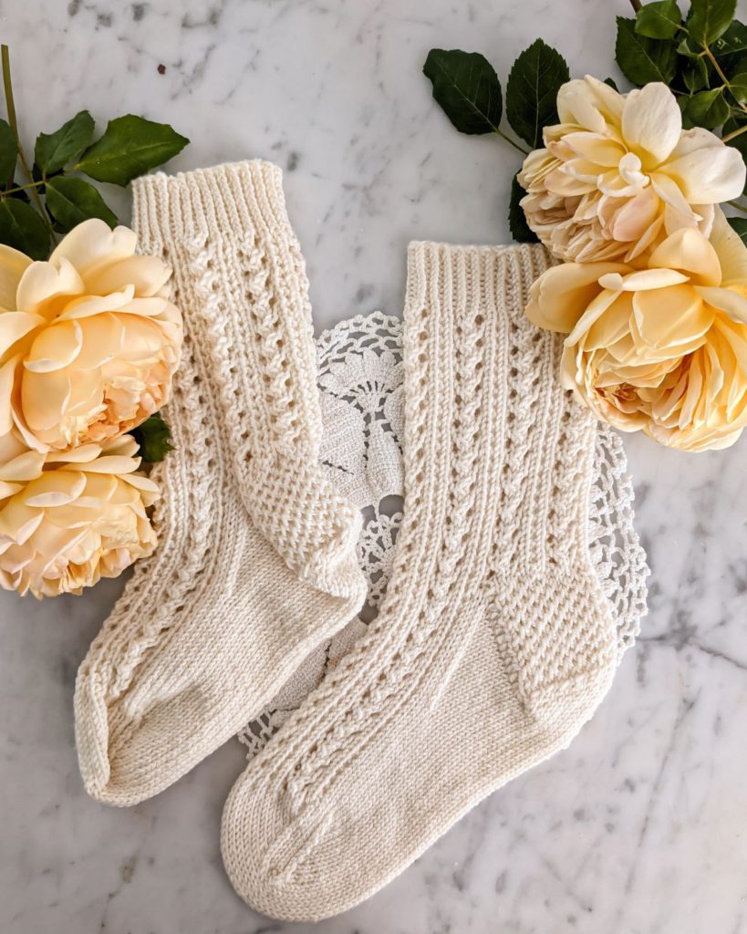 One blocked and one unblocked white sock are laid flat on a white marble countertop with yellow roses in the top corners. The unblocked sock looks lumpy, and shows really strong highlights and shadows. The blocked sock is smooth and the light hits it evenly. Blocking your project is a key step to getting better pictures of your knits.