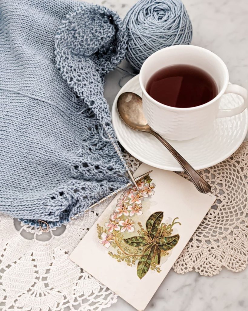 A partially finished, light blue sweater on metal needles is laid flat on a marble countertop next to a white teacup and saucer, some doilies, and an antique postcard with flowers on it. To edit your knitting photos, it helps to know what apps are available and which functions to use.