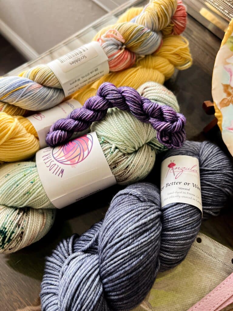 A pile of several full-sized skeins of yarn and a bright purple mini skein piled on a table.
