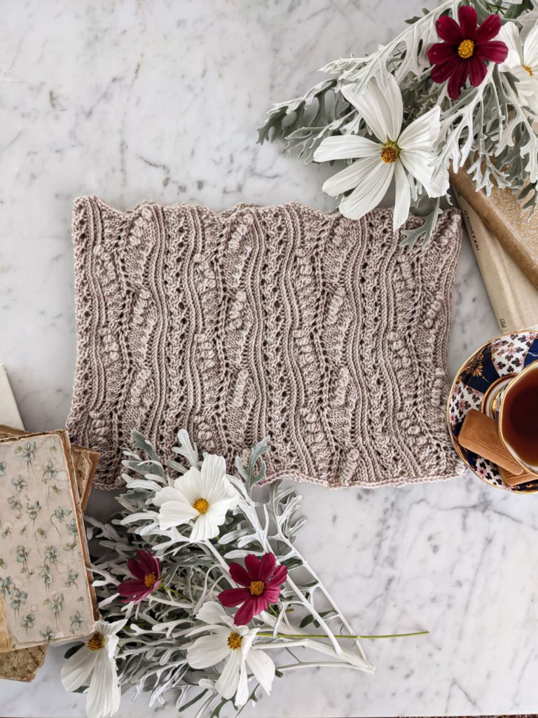 The Pandiculation Cowl, a DK-weight cowl with columns of tiny bobbles and eyelets, is laid flat on a white marble countertop surrounded by dusty miller, red and white cosmos, antique books, and a dark blue and white teacup.