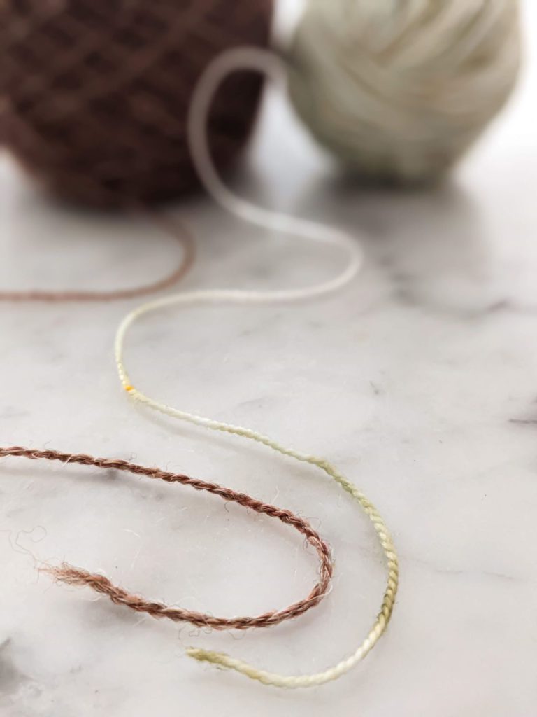 Two strands of yarn are splayed out on a white marble countertop. The yarn on the left, in brown, is woolen spun, resulting in a looser, fluffier yarn. The yarn on the right, in a pale green, is worsted spun, which makes a denser, tighter yarn. Understanding the difference between woolen spun and worsted spun is helpful when making yarn substitutions.