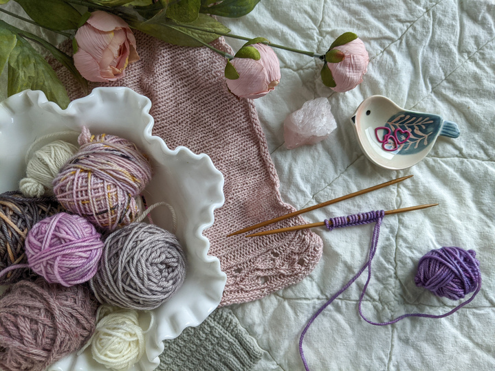 A bowl of soft purple, gray, and cream yarns sits on a white quilt along with a pink knit fabric swatch, some purple yarn on needles, and a little ceramic bird dish with stitch markers in it. When you're substituting yarn in your knit sweaters, swatching is crucial!